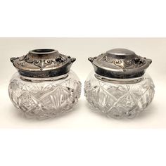 two glass jars are sitting next to each other on a white tablecloth, one has a silver lid and the other has an ornate design