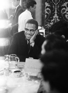 a black and white photo of a smiling man sitting at a table with people in the background