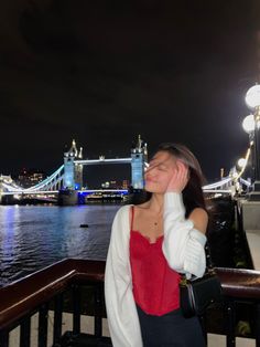 a woman standing next to the water in front of a bridge at night with her eyes closed