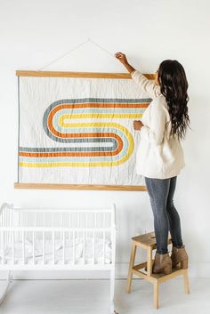 a woman hanging a quilt on the wall next to a baby crib and rocking chair