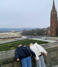 two people leaning against a stone wall with a clock tower in the background