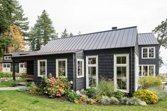 a small black house with lots of windows and flowers in the front yard, surrounded by greenery