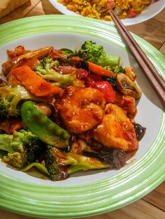 a green plate topped with chicken and veggies next to chopsticks on a wooden table