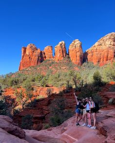 three women standing on the edge of a cliff in front of red rocks and trees