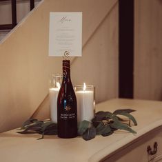 two candles and a bottle sitting on a table next to each other with a note in it