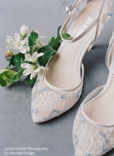 a pair of white wedding shoes next to a flower on a gray surface with green leaves