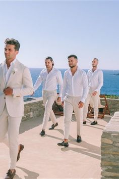 a group of men in white suits and ties are walking on the sidewalk by the ocean