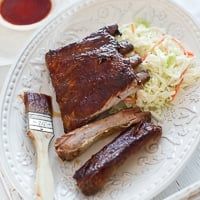 a white plate topped with ribs and coleslaw next to a cup of sauce