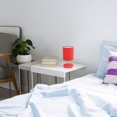 a white bed with blue sheets and pillows next to a table with a red lamp on it