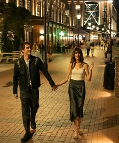 a man and woman holding hands walking down the street in front of buildings at night