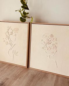 two framed art pieces with flowers on them next to a potted plant in the corner