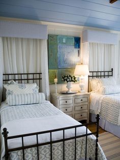 two beds in a room with white curtains and blue walls, one has a lamp on the nightstand