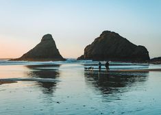 two people and their dog on the beach at sunset with rocks in the water behind them