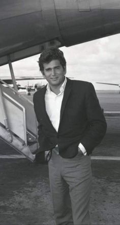 a man standing in front of an airplane with his hands in his pockets and looking at the camera