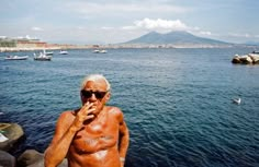 an older man standing on the edge of a body of water with boats in the background