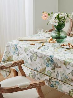 a table with flowers and plates on it in front of a chair next to a window