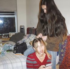 two women are sitting on the floor and one is looking at her cell phone while another woman stands nearby