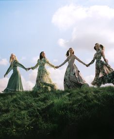 four women in long dresses holding hands while standing on top of a grass covered hill