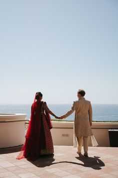 the bride and groom are holding hands as they stand on a balcony overlooking the ocean