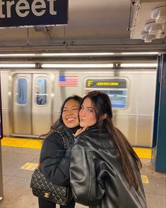 two young women hugging each other in front of a subway train at the station with their arms around each other