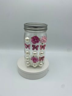 a glass jar with pink flowers and pearls on the lid is sitting on a white base