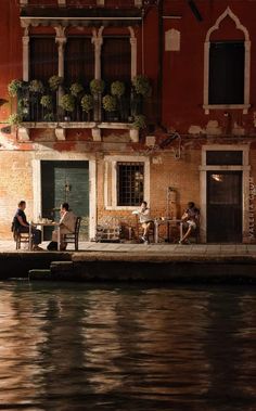 some people sitting at tables on the side of a building next to a body of water