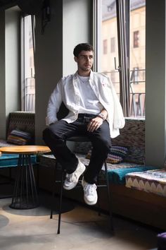 a man sitting on a chair in front of a window with his legs crossed and wearing white sneakers