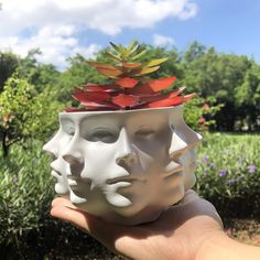 a hand holding a white ceramic planter with three faces on it's face