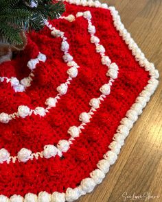 a red and white crocheted christmas tree skirt on a wooden floor next to a fir tree