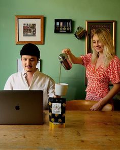 a man and woman sitting at a table in front of a laptop computer, pouring coffee into a cup