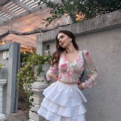 a woman standing on the sidewalk in front of a building wearing a white skirt and pink top