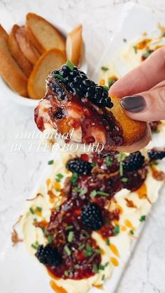 a person holding up a piece of bread with berries on it and other food items in the background