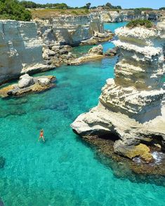 a person swimming in the blue water near some rocks and cliffs with green vegetation on either side