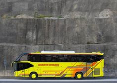 a yellow bus driving down the road next to a large rock wall and grass area