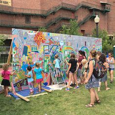 several people are painting on a large wall in the grass near a brick building with stairs