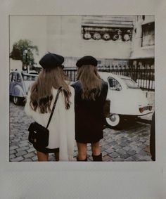 two women standing next to each other in front of a white car on a cobblestone street