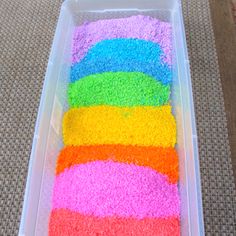 there is a plastic container filled with rice and rainbow colored rice on the floor next to it