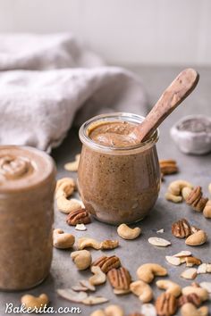 a jar filled with peanut butter surrounded by nuts on a table next to a wooden spoon