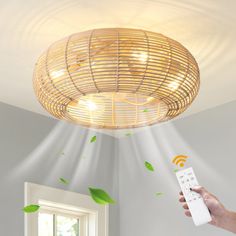 a hand holding a remote control in front of a wicker light fixture with green leaves on the ceiling