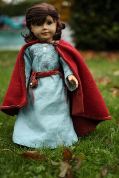 a doll in a blue dress and red cape is standing in the grass with her hands on her hips