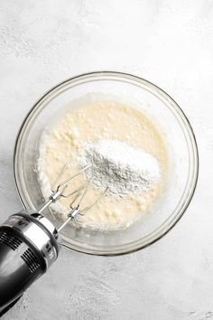 a whisk and flour in a glass bowl on a white table with an electric hand mixer