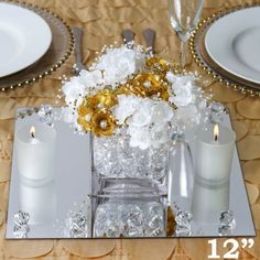 the table is set with white plates and silverware, gold flowers and candlesticks