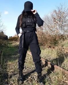 a woman in black is standing on railroad tracks wearing all black clothing and a hat