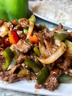 a white plate topped with beef and peppers next to rice on a tablecloth covered table