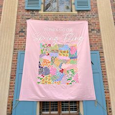 a pink banner hanging from the side of a brick building with blue shutters and windows