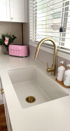 a white kitchen sink sitting under a window next to a pink vase with flowers on it