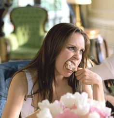 a woman sitting in front of a cake with frosting on it and eating something out of her mouth