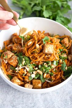 a person is holding a spoon over a bowl of noodles with carrots and greens