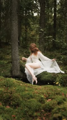 a woman in white dress sitting on mossy ground next to a tree with her legs spread out