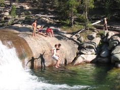 some people are swimming in the water near rocks and trees, while another person is standing on top of a large rock